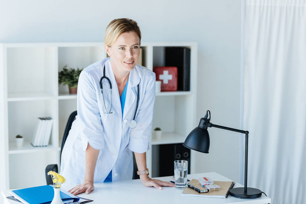 ελκυστική γυναίκα γιατρό σε λευκό παλτό με στηθοσκόπιο πάνω από λαιμό μιλώντας στο γραφείο  - Φωτογραφία, εικόνα