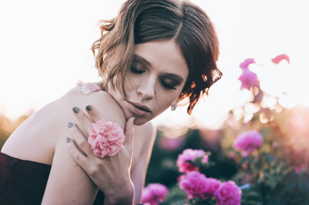 Hermosa joven con el pelo rizado posando cerca de rosas en un jardín. El concepto de publicidad de perfumes
. - Foto, imagen