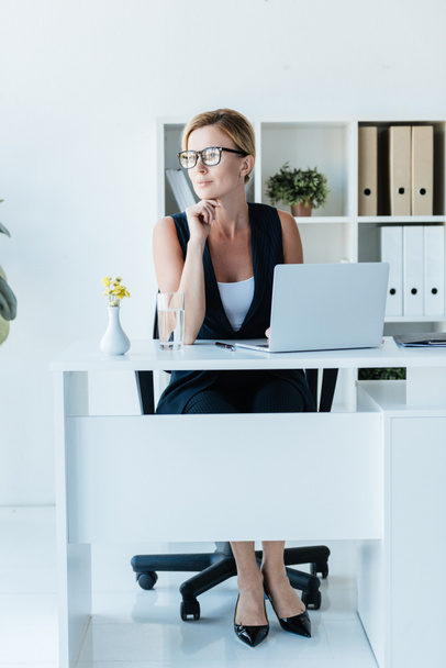 mujer de negocios adulta pensativa en anteojos sentados en la mesa con el ordenador portátil en la oficina
  - Foto, Imagen