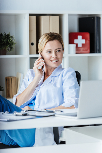 female doctor looking away and talking on smartphone at table with laptop in office - Foto, Bild