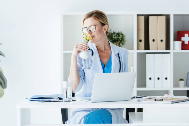adult female doctor in eyeglasses smelling flowers at table with laptop in office  - Foto, afbeelding
