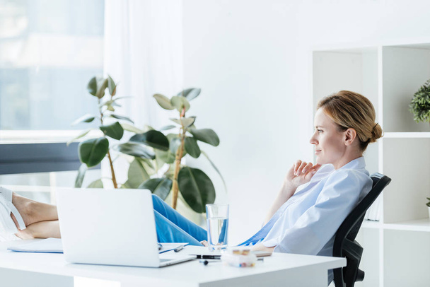 side view of thoughtful female doctor with legs on table sitting in office  - Foto, imagen