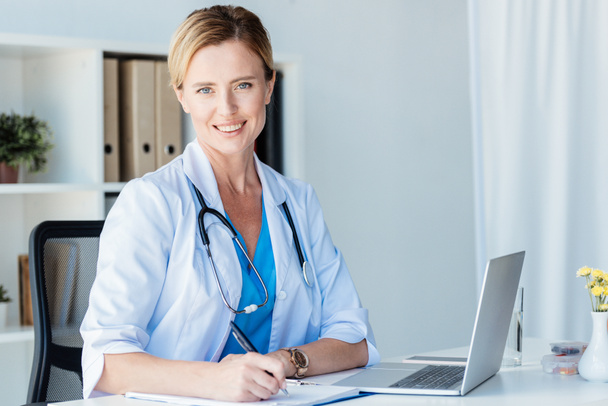 smiling female doctor looking at camera while writing in clipboard at table with laptop in office  - Photo, Image