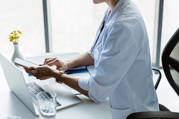 vue partielle du médecin féminin à l'aide d'un smartphone à la table avec ordinateur portable au bureau
  - Photo, image