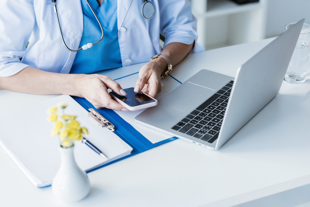 image recadrée de femme médecin à l'aide d'un smartphone avec écran blanc à la table avec ordinateur portable au bureau
  - Photo, image