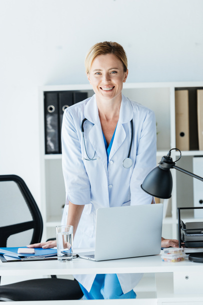 ευτυχισμένη γυναίκα γιατρό σε λευκό παλτό με στηθοσκόπιο πάνω λαιμό στέκεται στο τραπέζι με το laptop στο γραφείο  - Φωτογραφία, εικόνα