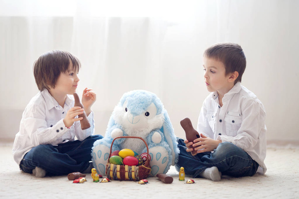 Two adorable little children, boy brothers, having fun eating chocolate bunnies and playing with eggs at home, back light - Фото, изображение