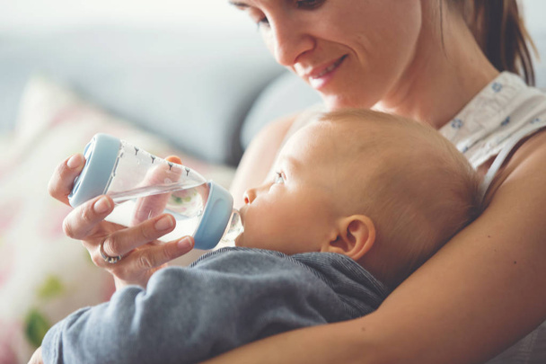 Mother, feeding her baby boy from bottle, comfortably sitting on the couch at home - Photo, Image