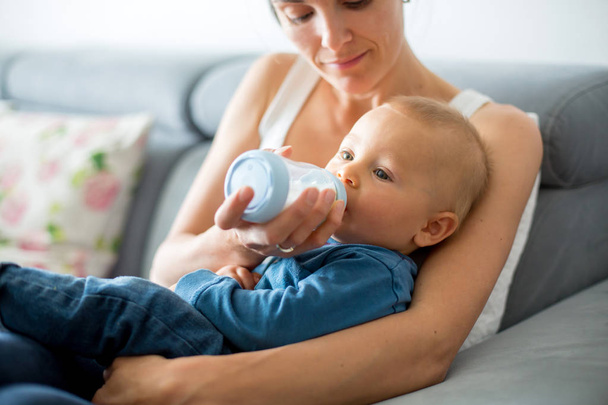 Mother, feeding her baby boy from bottle, comfortably sitting on the couch at home - Photo, image