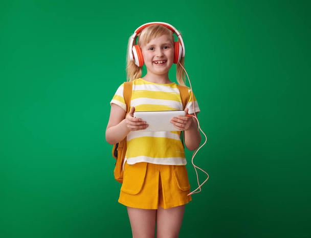 smiling pupil with backpack holding tablet PC and listening to music on green background - Foto, Imagen