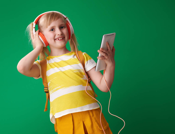 smiling school girl with backpack holding tablet PC and listening to music in headphones on green background  - Фото, изображение