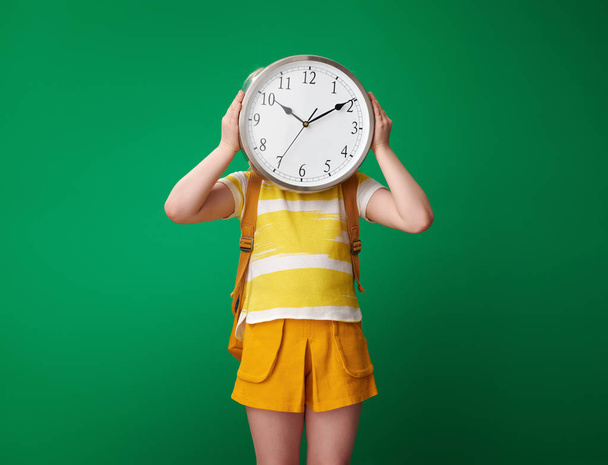 pupil with backpack holding clock in front of face against green background - Foto, immagini