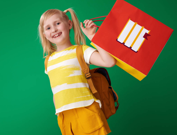 happy pupil with backpack holding shopping bag with stationery on green background - Fotografie, Obrázek