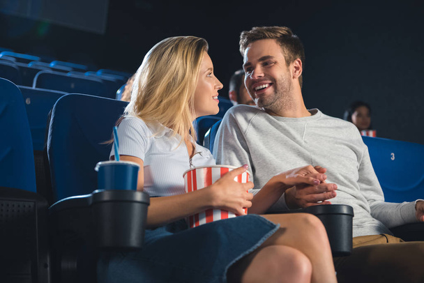 smiling couple with popcorn and soda drink holding hands while watching film together in cinema - Foto, Imagem