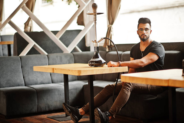Stylish beard arabian man in glasses and black t-shirt smoking hookah at street bar. Arab model having rest. - Foto, imagen