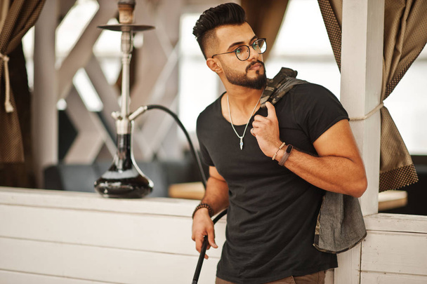 Stylish beard arabian man in glasses and black t-shirt smoking hookah outdoor. Arab model having rest. - Fotografie, Obrázek