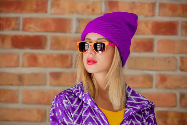 Young blonde woman in 90s sports jacket and hat on brick wall background - Photo, Image