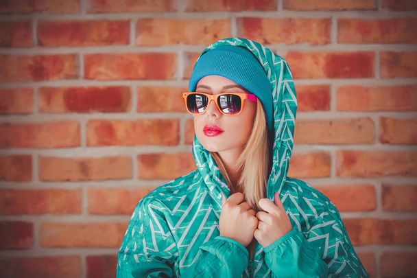 Young blonde woman in 90s sports jacket and hat on brick wall background - Photo, Image