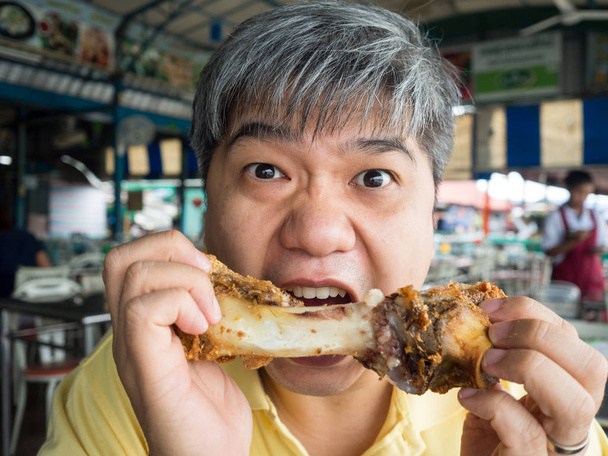 Ασιατικές άνθρωπος τρώει χοιρινό κόκκαλο μετά το φαγητό στο πόδι της τραγανό ψημένο στη σχάρα χοιρινό κότσι. Στο εστιατόριο του ποταμού στην Ταϊλάνδη. Αυτός φαίνεται νόστιμο και απολαμβάνει να τρώει τόσο πολύ. - Φωτογραφία, εικόνα