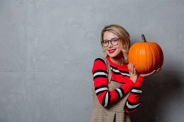 Portrait of a young stylish blonde girl in red striped sweater with halloween pumpkin on grey background. - Fotografie, Obrázek