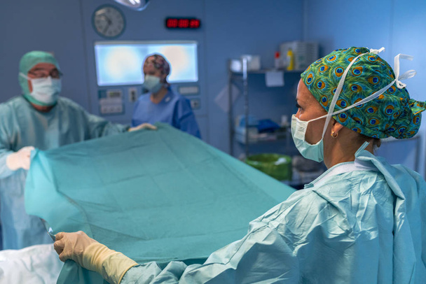 Χειρουργός και βοηθός τοποθετώντας το αποστειρωμένο φύλλο πάνω από τον ασθενή. Νοσοκόμα σε την πίσω περιμένοντας. - Φωτογραφία, εικόνα