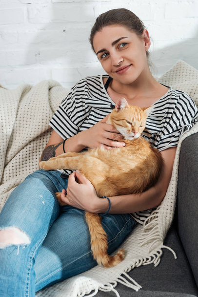 beautiful smiling young woman holding red cat and sitting on couch - Valokuva, kuva