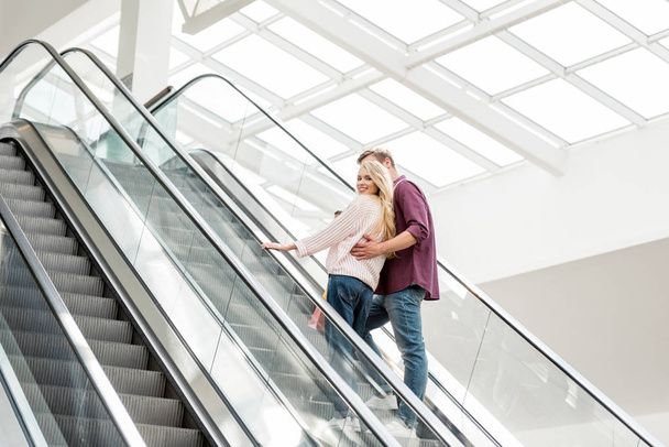 vista trasera de la joven pareja de compradores con bolsas de papel en la escalera mecánica en el centro comercial
  - Foto, Imagen