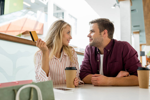couple with credit card and disposable cups of coffee looking at each other at table in cafe  - Foto, Imagem