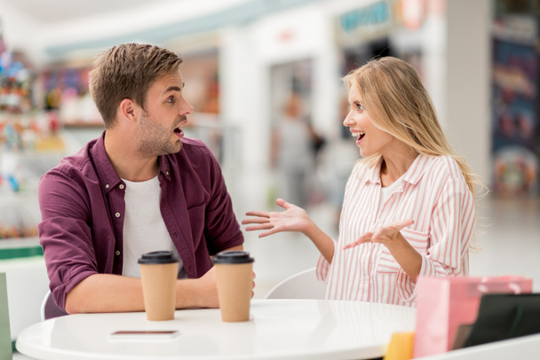shocked young woman gesturing by hands and looking at boyfriend while he sitting near at table with coffee cups in cafe - Photo, Image