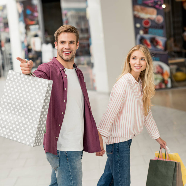 handsome young man with shopping bags pointing to girlfriend walking near at shopping mall - Photo, Image