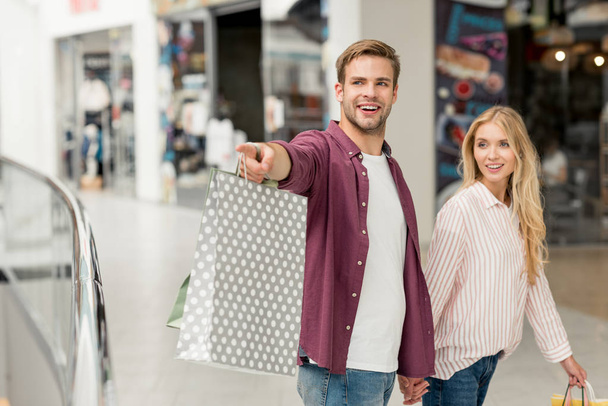 foyer sélectif de sourire jeune homme avec des sacs à provisions pointant vers petite amie marchant près au centre commercial
 - Photo, image