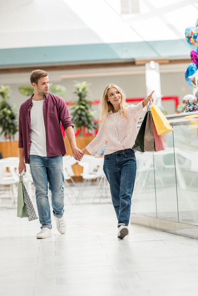 belle jeune femme avec des sacs à provisions pointant vers petit ami marchant près au centre commercial
 - Photo, image