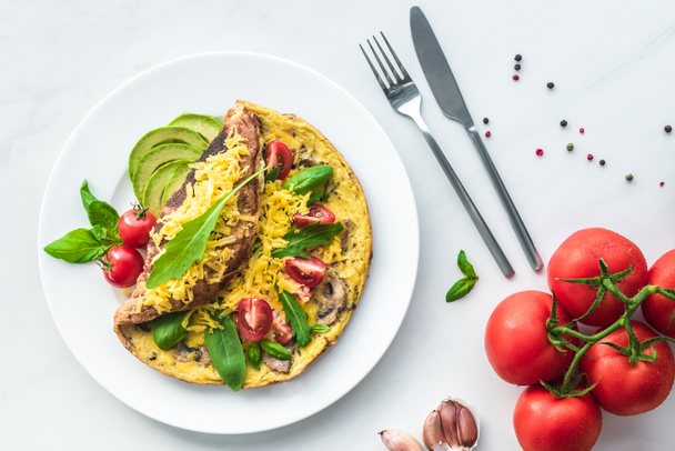 flat lay with omelette with cherry tomatoes, avocado pieces and cutlery on white marble surface - Фото, изображение