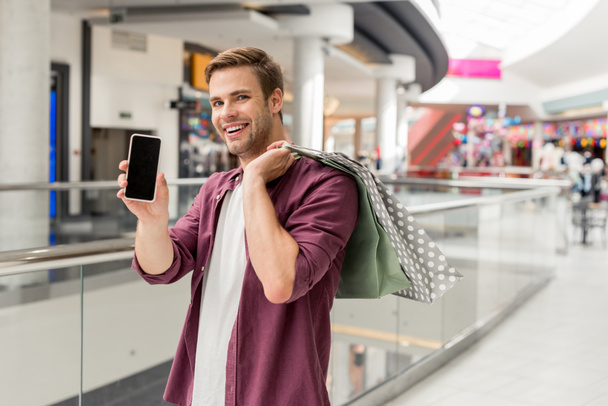 sourire jeune homme avec des sacs à provisions montrant smartphone avec écran blanc au centre commercial
  - Photo, image