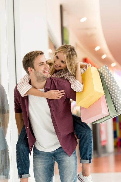 smiling man giving piggyback ride to girlfriend with shopping bags at mall  - 写真・画像