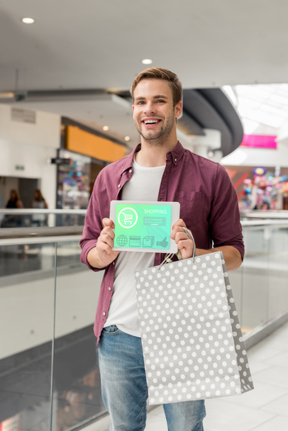 smiling man with paper bag showing digital tablet with shopping on screen at mall  - Foto, Bild