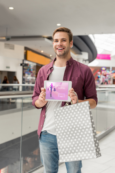 молодий красивий чоловік з паперовим мішком, що показує цифровий планшет з онлайн-покупками на екрані в торговому центрі
  - Фото, зображення