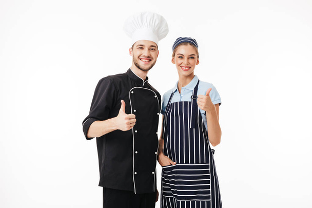 Jeune homme chef souriant en uniforme noir et chapeau blanc et jolie femme cuisinier en tablier rayé et casquette regardant joyeusement à la caméra montrant de gros pouces sur fond blanc
 - Photo, image