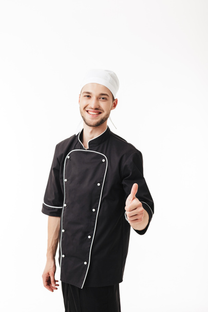 Jovem homem sorridente chef em uniforme preto alegremente olhando na câmera mostrando polegar acima gesto sobre fundo branco
 - Foto, Imagem