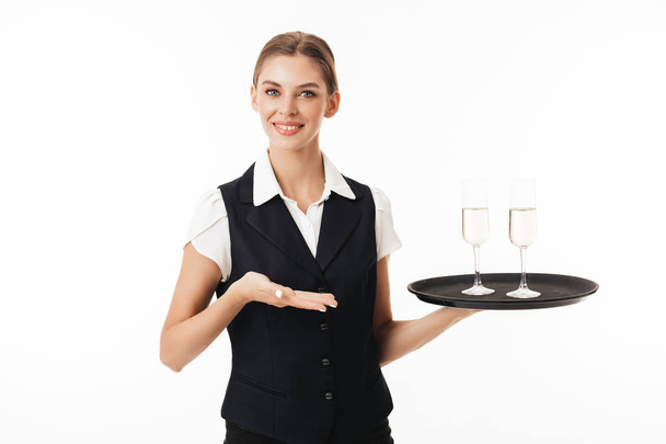 Young smiling waitress in uniform holding tray with glasses joyfully looking in camera over white background - Photo, Image