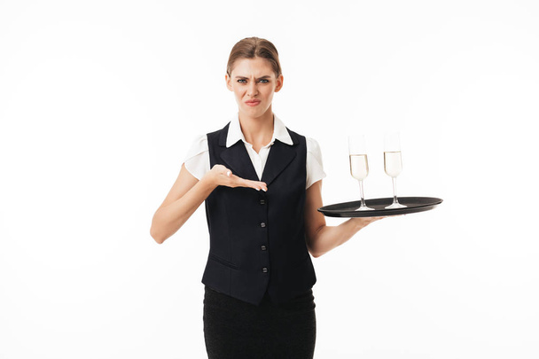 junge Kellnerin in Uniform hält Tablett mit Gläsern und blickt traurig in die Kamera über weißem Hintergrund - Foto, Bild