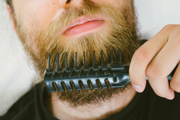 Bearded man taking care of his facial hair, brushing beard using brush comb. - Foto, afbeelding