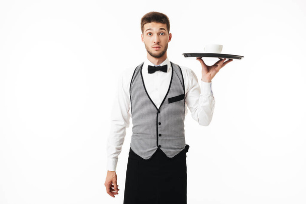 Joven camarero sorprendido en bandeja de sujeción uniforme con taza de café asombradamente mirando en la cámara sobre fondo blanco
 - Foto, imagen