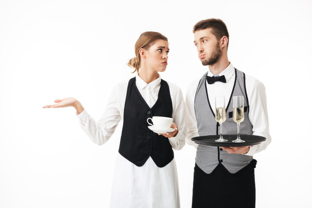 Young waiter holding tray with glasses of champagne and sad waitress holding cup of coffee in hands tiredly looking at each other over white background - Valokuva, kuva