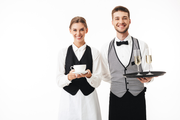 Młody kelner wesoły taca z kieliszków szampana i dość kelnerka filiżankę kawy w rękach szczęśliwie w aparacie razem patrząc na białym tle - Zdjęcie, obraz