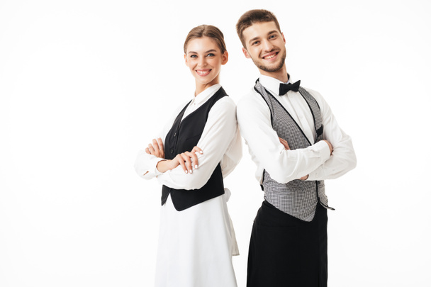Young smiling waiter and waitress in white shirts and vests sstanding back to back joyfully looking in camera with arms folded over white background - Photo, Image