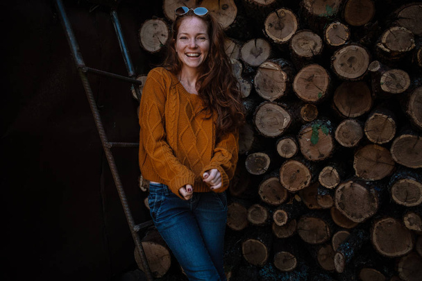 beautiful amazing gorgeous redhair girl spins in yellow sweater, jeans and glasses on the background of firewood. Autumn mood - Фото, изображение