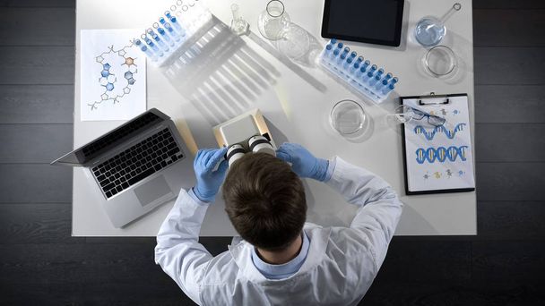 Vista superior del trabajador de laboratorio examinando la muestra preparada bajo el microscopio
 - Foto, imagen