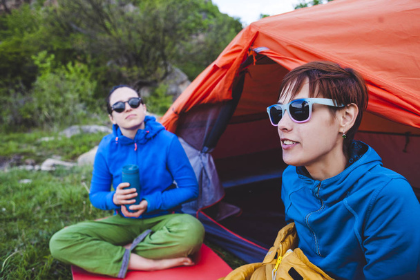 Due ragazze sono sedute in una tenda. Le fidanzate comunicano in natura. Le donne nei sacchi a pelo bevono caffè. Amici nel campeggio. Brune sorridenti parlando
. - Foto, immagini