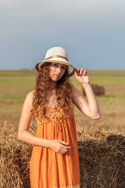 vertical portrait of a young cute rural woman in an orange sarafan and a hat near a stack of wheat hay on a field in the rays of the setting sun - Fotó, kép